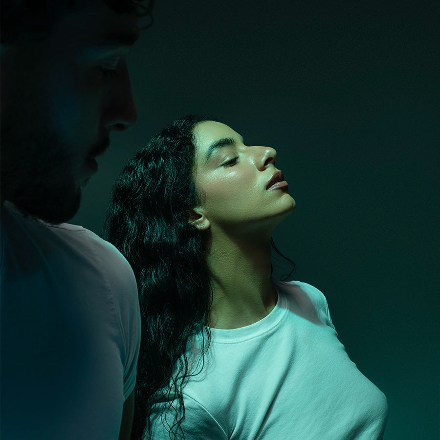 A woman leans back into green-tinted light, eyes closed, while a figure stands just behind her in shadow. An intimate, cinematic portrait echoing the atmospheric, emotional world of Looking for Each Other fragrances.