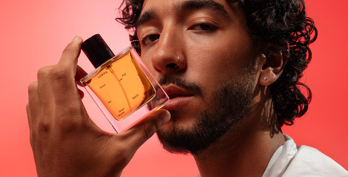 A model holds a 50ml bottle of Quasi Life in the Quasi World by Looking for Each Other against a bold coral-red backdrop. The golden botanical fragrance glows in the glass bottle, capturing the brand’s genderless, natural approach to scent—made without synthetics or isolates.
