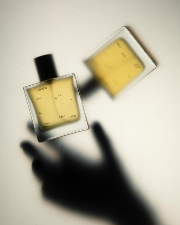 Two bottles of Looking for Each Other botanical fragrance cast soft shadows on a cream surface, with a dark hand silhouette reaching toward the glass. A poetic still life that reflects the brand’s genderless, botanical fragrance philosophy—made without synthetics or isolates.