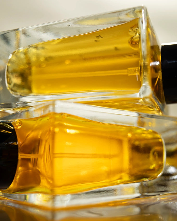 Close-up of two Looking for Each Other perfume bottles stacked on their sides, glowing with golden botanical liquid. A detailed look at the richness of ingredients inside—100% natural, genderless, and crafted from essential oils in organic grape alcohol.