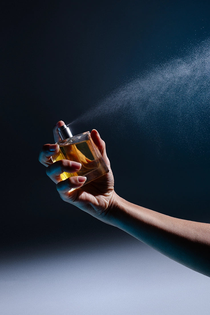 A hand sprays a bottle of botanical fragrance from Looking for Each Other into a mist of fine vapor against a deep navy background. The amber liquid glows through the glass, capturing the purity and performance of scent made without synthetics.