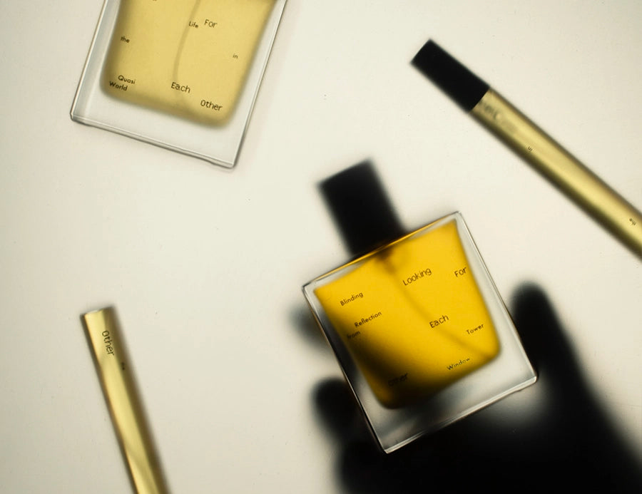 Flatlay of Looking for Each Other’s two signature fragrances—Quasi Life in the Quasi World and Blinding Reflection from Tower Window—alongside two 10ml-size vials. The golden botanical liquid glows under soft light as a hand’s shadow reaches toward the bottle, evoking intimacy and the brand’s minimal, poetic ethos.